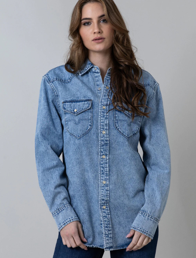 A female model wearing a blue oversized boyfriend button-down denim shirt with two front pockets and pearl snaps.