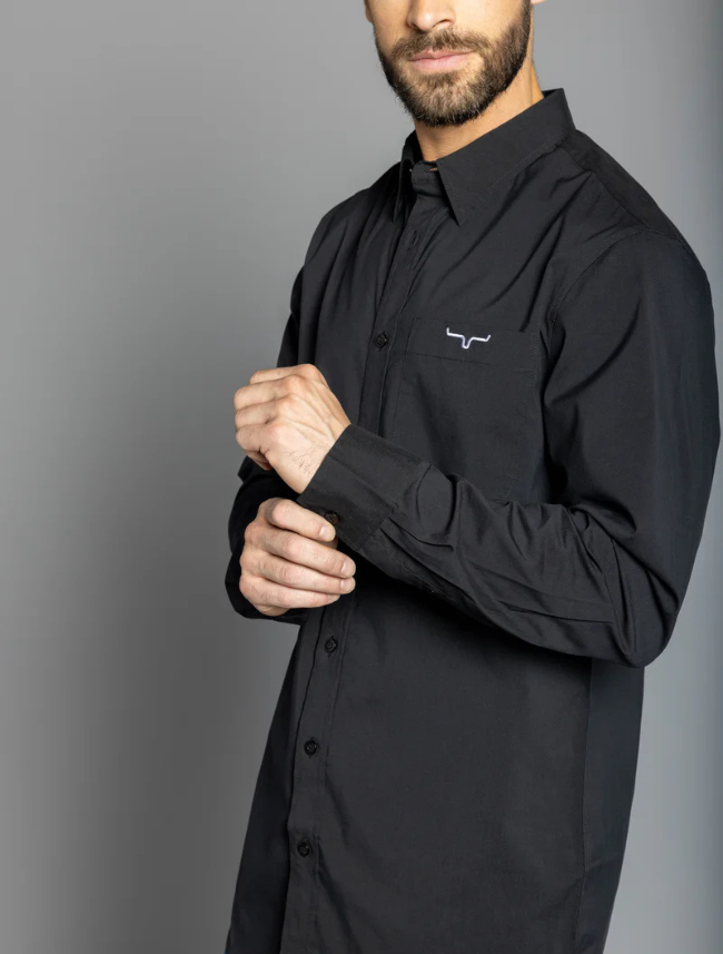 A man wearing a long-sleeved black dress shirt with a single chest pocket and a small embroidered logo on the chest area. The shirt has a button-up front and is modeled in a standing position against a gray background.