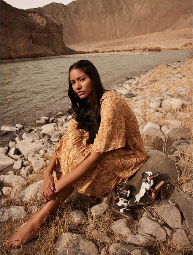 Woman in a gold dress sitting on rocks by a river with mountains in the background with a pair of cowhide and brown leather booties