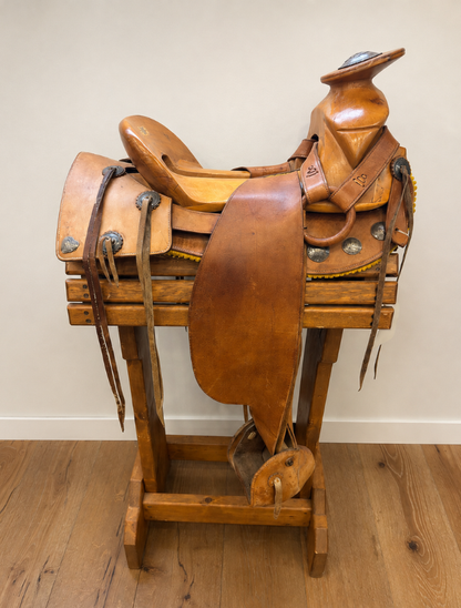 A brown leather western saddle displayed on a wooden stand in an indoor setting with a beige wall and wood floor.
