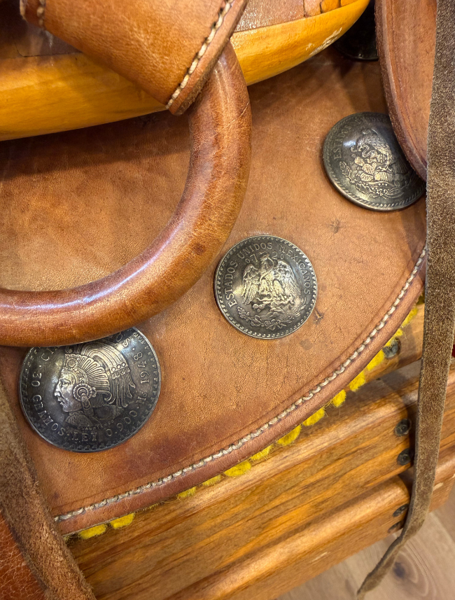 Close-up of a brown leather saddle with large silver coin medallions and a curved leather handle.