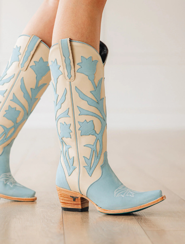 Light blue and beige cowboy boots with floral patterns on a light wooden floor.