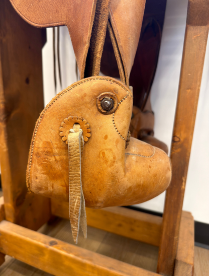 A close-up of a vintage tan leather saddle foothold with a decorative strap and metal snap, hanging on a wooden display stand.