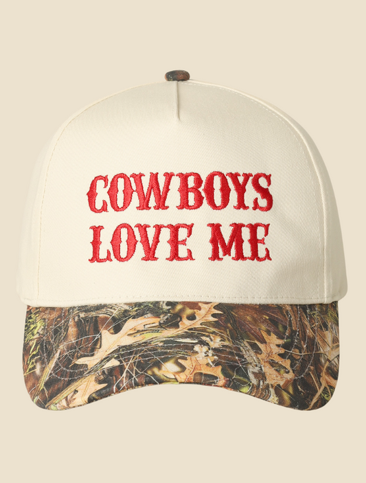 Trucker hat with “Cowboys Love Me” embroidered in bold red letters on a structured white front panel with a camo brim.