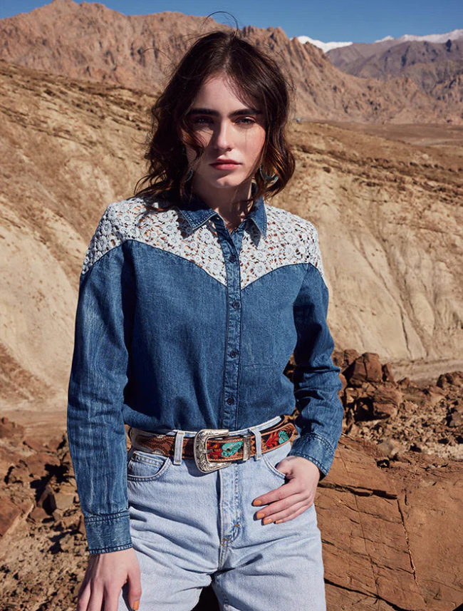 Woman wearing a blue denim shirt with white patterned collar and light-wash denim jeans with a multicolor floral tooled leather belt in a desert landscape