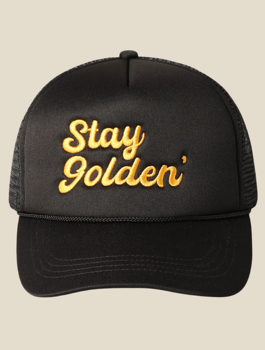 Black trucker hat with “Stay Golden Cowgirl” embroidered in metallic gold thread on the front panel, featuring mesh back and curved brim.