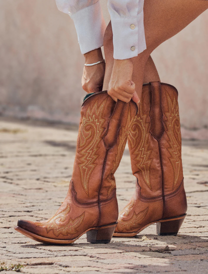 Brown cowboy boots with intricate designs worn by a person on a stone path.